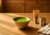 Ceramic bowl of vibrant green matcha latte with bamboo whisk and matcha powder tin on a wooden counter — matcha tea benefits