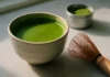 Matcha latte keraminiame puodelyje ant balto marmuro su bambukiniu šluotele ir matcha miltelių dėžute